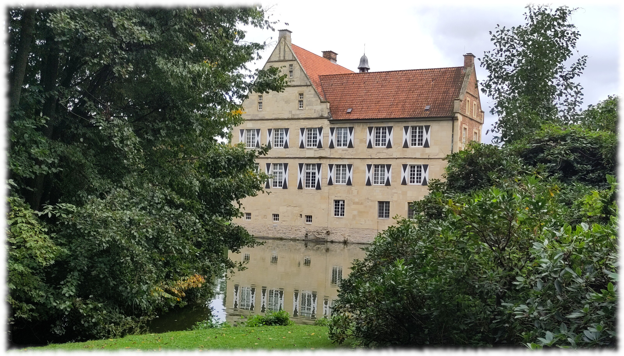Burg Hülshoff mit Spiegelung im Wasser des Burggrabens, eingerahmt von Bäumen und Sträuchern. Himmel bedeckt. 