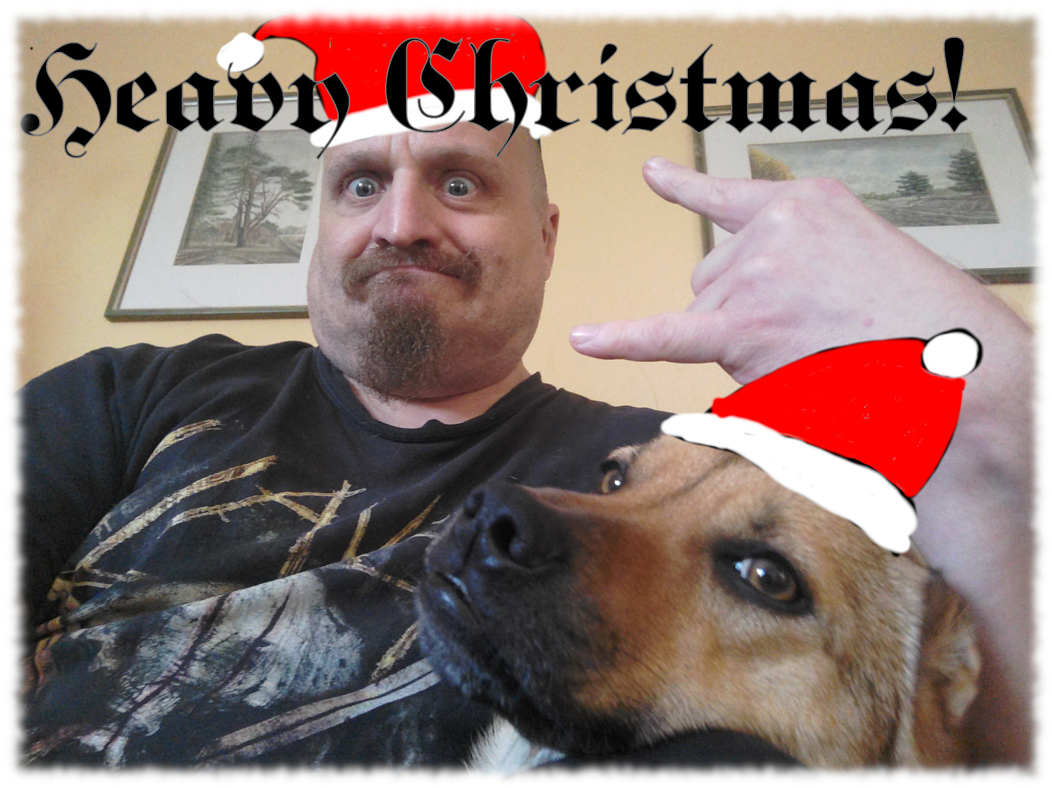 Ulf mit Ziegenbart und schwarzem Slayer-T-Shirt zeigt mit der rechten Hand das Metal-Gru�zeichen, dar�ber der Text 'Heavy Christmas!' in schwarzer Schrift Auf dem Scho� ist der Kopf der braunen H�ndin Trixi zu sehen. Bei beiden ist eine Weihnachtsmannm�tze auf den Kopf gemalt